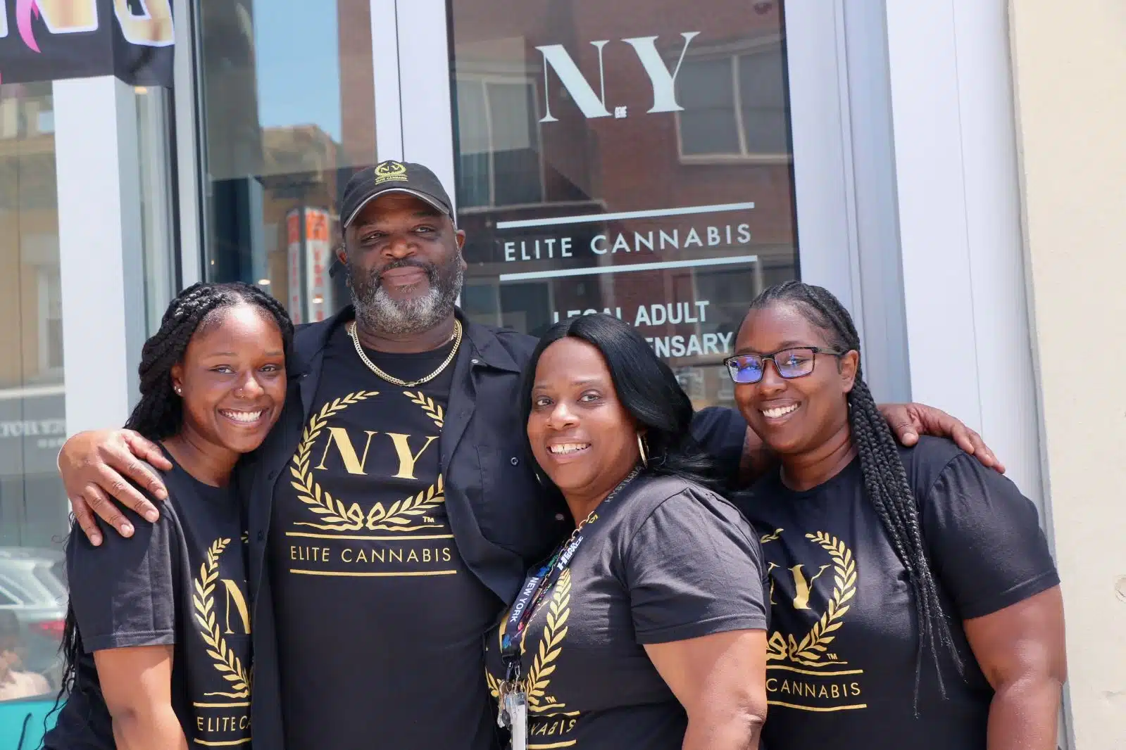 Carson Grant and the NY Elite Cannabis family team in front of the Bayside storefront, wearing the gold-laurel logo on black tees
